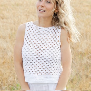 Pode incluir: Top de crochê branco com um padrão quadrado. O top sem mangas tem decote redondo e bainha canelada. O fundo é um campo de grama seca.