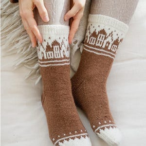 Könnte beinhalten: Ein Paar braune und weiße Strickstrümpfe. Die Socken haben einen weißen Bund mit einem sich wiederholenden Muster von Gebäuden. Der Fuß- und Beinbereich der Socken ist braun. Die Socken sind aus einem weichen, warmen Material.