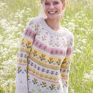 May include: A cream-colored sweater with a floral pattern in pink, yellow, and green. The sweater has a round neckline and long sleeves. The floral design includes tulips and other flowers. The background is a field of white flowers.