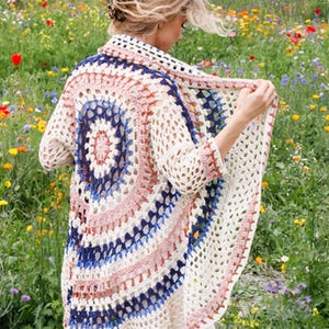 May include: A crochet cardigan featuring a circular mandala pattern on the back. The garment is crafted with off-white, navy blue, and dusty pink yarn, showcasing an openwork stitch design with three-quarter length sleeves and a draped open front.