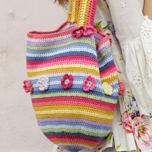 Op de afbeelding: Een handgemaakte gehaakte tote bag met horizontale strepen in verschillende kleuren, waaronder roze, geel, blauw en groen. De tas is versierd met kleine, driedimensionale gehaakte bloemen in roze en geel. De tas heeft een afgeronde vorm en twee handvatten.
