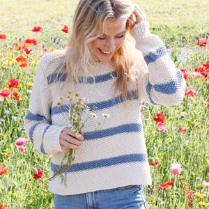 May include: Cream-colored sweater with blue horizontal stripes on the body and sleeves. The sweater is made of a textured knit fabric. The person is holding a small bouquet of white and yellow flowers in a field of colorful wildflowers.