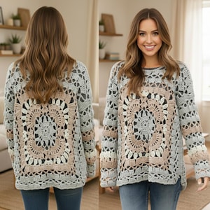 Op de afbeelding: Een lichtgrijze gehaakte trui met een vierkant, bloemachtig ontwerp in het midden, met lichtbeige en grijze accenten. De trui met lange mouwen heeft een ronde halslijn en wordt gedragen met een blauwe spijkerbroek.