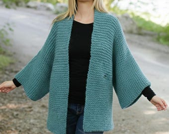 May include: A teal-colored, open-front cardigan with wide sleeves. The knitted sweater has a textured appearance and a relaxed fit. The cardigan is worn over a black top and blue jeans. The sleeves are long and wide, and the cardigan has a mid-thigh length.