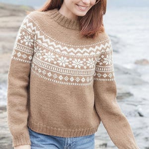 Könnte beinhalten: Ein beigefarbener Langarmpullover mit einem weißen Fair-Isle-Muster an der Passe, der Brust und den oberen Ärmeln. Das Kleidungsstück hat einen gerippten Stehkragen, gerippte Bündchen und einen gerippten Saum, kombiniert mit einer blauen Jeans.