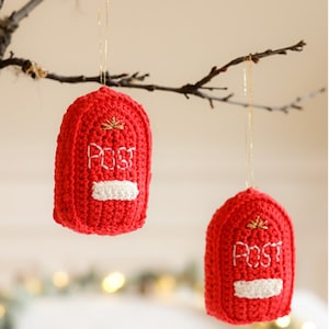 Puede incluir: Dos adornos rojos de ganchillo con forma de buzones británicos, colgando de una rama. Cada adorno tiene la palabra "Post" bordada en blanco, con un detalle rectangular blanco. Hilo dorado para colgar.