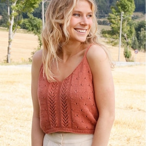 Puede incluir: Un top de camisola de punto color coral con cuello en V y delicadas correas. El top presenta un patrón de punto detallado con detalles calados. La modelo lleva el top con pantalones de color claro.