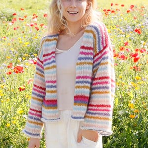 Op de afbeelding: Een kleurrijk gestreept vest in roze, rood, blauw en geel. De open trui is gebreid met een getextureerde steek. Het vest wordt gedragen over een witte top. De achtergrond is een veld met wilde bloemen.