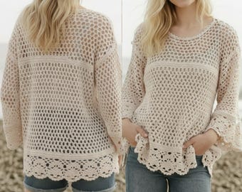 May include: A light beige crochet sweater with a round neckline and long sleeves. The sweater features a loose, open weave design with a decorative scalloped hemline. The model is wearing denim shorts.