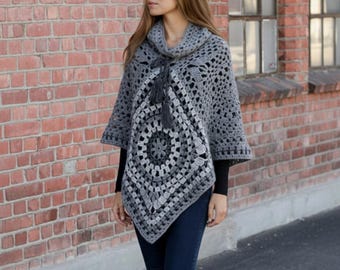 Puede incluir: Un poncho de ganchillo gris con cuello alto y diseño geométrico. El poncho presenta un patrón circular central y un detalle de borla en el escote. La prenda se lleva sobre una camiseta negra de manga larga y vaqueros azul oscuro.