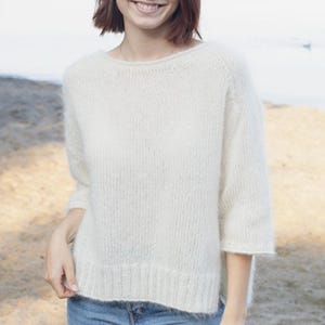 May include: Off-white, crew neck sweater with three-quarter sleeves. The sweater has a ribbed hem and cuffs. The knitwear appears soft and fluffy. The model is wearing blue jeans.