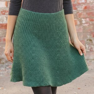 Könnte beinhalten: Ein grüner Strickrock mit ausgestellter A-Linien-Silhouette. Der Rock hat vertikale Rippen und dekorative Details. Der Rock hat eine einfarbige, mittelgrüne Farbe und scheint aus einem weichen, warmen Material zu bestehen.