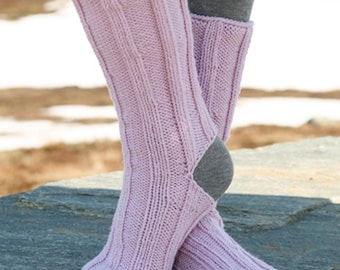 May include: A pair of light pink knitted leg warmers with a ribbed design. The leg warmers are worn over gray socks and are resting on a stone surface. The knitted leg warmers are a soft, pastel color.