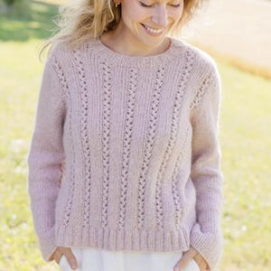 Puede incluir: Un suéter de punto rosa claro con cuello redondo y mangas largas. El suéter presenta detalles calados verticales y un dobladillo y puños acanalados. La prenda de punto suave y texturizada es adecuada para climas más fríos.