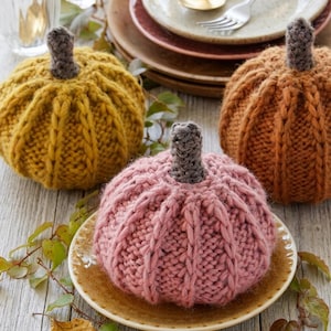 May include: Three hand-knitted pumpkins in mustard yellow, dusty pink, and burnt orange. Each pumpkin features a thick, ribbed knit texture and a small, dark grey crocheted stem. The pumpkins are arranged on a light-coloured wooden surface.