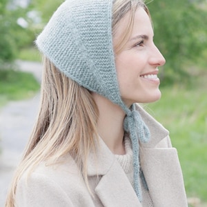 May include: A light sage green knit bonnet featuring a textured garter stitch pattern and long matching ties that fasten under the chin. The headwear is crafted from a soft, fuzzy yarn and covers the top and back of the head securely.