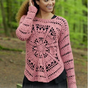 Puede incluir: Un suéter de crochet rosa polvoriento con cuello redondo y mangas largas. El suéter presenta un diseño calado con un patrón circular en la parte delantera y detalles a rayas en las mangas. El dobladillo inferior es curvo.