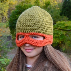 May include: A crochet beanie hat featuring an olive green crown and a bright orange mask-style band across the eyes. The hat is constructed with a dense, textured stitch pattern, creating a fitted silhouette that covers the top of the head.