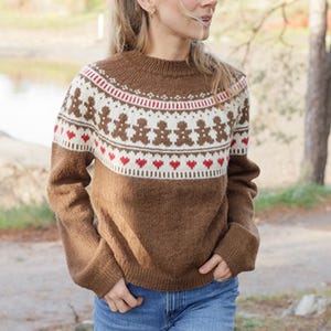 May include: Brown sweater with a round neck and long sleeves. The sweater features a white band with red accents and gingerbread man figures. Below the band is a row of red hearts. The model is wearing blue jeans.