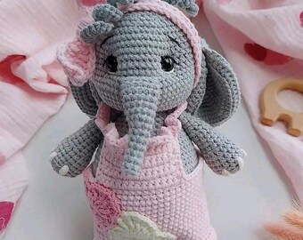 Puede incluir: Una muñeca elefante de ganchillo hecha a mano en gris y rosa. El elefante lleva un peto rosa con una aplicación floral y una diadema a juego. La muñeca tiene ojos negros y una larga trompa. El fondo incluye tela rosa.