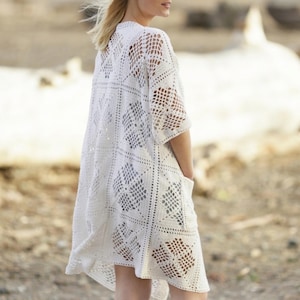 May include: A white, open-weave crochet cardigan with short sleeves and pockets. The cardigan features a geometric pattern throughout the design. The model is wearing the cardigan outdoors.