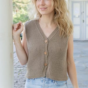 Puede incluir: Un chaleco sin mangas de punto beige con cuello en V y botones de madera. El chaleco está abotonado y tiene una textura acanalada. La modelo lleva vaqueros azules.