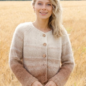 May include: A button-down cardigan in a gradient of cream to brown. The long-sleeved sweater has a round neckline and is made of a soft, textured yarn. The cardigan is worn over a white garment, with a field of wheat in the background.