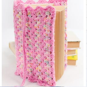 May include: A pink crocheted book cover with a scalloped edge is wrapped around a book. The cover has a textured pattern with flecks of color. The book's pages are visible, and other books are stacked in the background.
