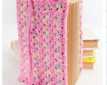 May include: A pink crocheted book cover with a scalloped edge is wrapped around a book. The cover has a textured pattern with flecks of colour. The book's pages are visible, and other books are stacked in the background.