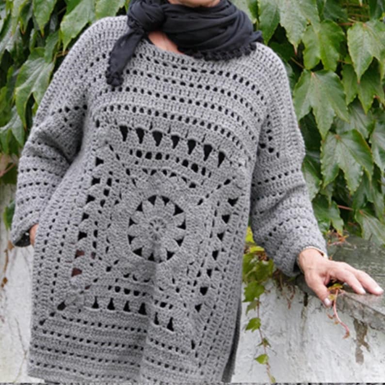 May include: A gray crochet tunic with long sleeves and a decorative square pattern on the front. The tunic has a round neckline and a loose fit. The garment is made of a soft, textured yarn and is suitable for layering.
