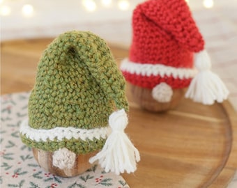May include: Two handmade gnome ornaments. One has a green knitted hat with a white tassel and trim, the other has a red knitted hat with a white tassel and trim. Both have wooden bodies and are on a wooden tray.