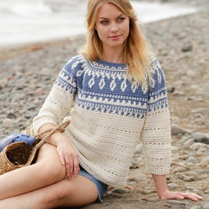 May include: A cream-colored crochet sweater with blue and white geometric patterns on the yoke. The sweater has three-quarter sleeves and a round neckline. The model is sitting on a sandy beach, wearing denim shorts and holding a straw bag.