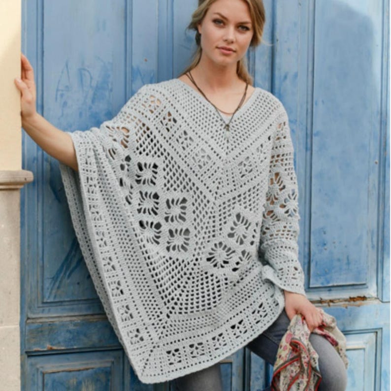 Op de afbeelding: Een lichtgrijze, open geweven gehaakte poncho met een V-hals. De poncho heeft een bloemenpatroon en lange mouwen. De vrouw draagt de poncho met een jeans en een ketting.