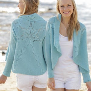 Könnte beinhalten: Hellblauer Strickcardigan mit floralem Design auf dem Rücken. Der Cardigan hat lange Ärmel und eine drapierte Front. Das Model trägt weiße Shorts. Der Hintergrund ist eine Strandszene.