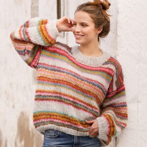 Puede incluir: Un suéter holgado y acogedor con rayas horizontales multicolores, incluyendo rosa, naranja, amarillo y azul. El suéter tiene cuello redondo y mangas largas. El fondo es una pared blanca.
