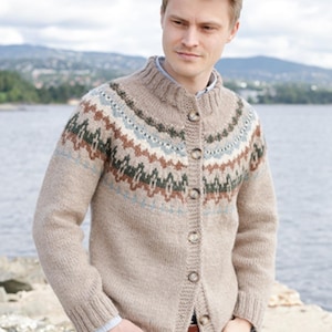Op de afbeelding: Een beige gebreid vest met een ronde hals en een knoopsluiting aan de voorzijde. De bovenkant van de borst en de schouders zijn versierd met een horizontale band van geometrische patronen in tinten donkergroen, lichtblauw en bruin.