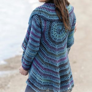 Op de afbeelding: Een handgemaakt, open geweven gehaakt vest in tinten blauw, paars en grijs. De achterkant heeft een cirkelvormig ontwerp en de mouwen zijn lang. Het vest is een uniek, draagbaar kunstwerk.