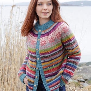 Könnte beinhalten: Ein bunter Cardigan mit Knöpfen und rundem Ausschnitt. Der Pullover hat horizontale Streifen in verschiedenen Farben, darunter Rot, Blau, Grün und Lila. Schneeflockenmuster sind auf der oberen Brust zu sehen. Der Cardigan wird mit einer blauen Jeans getragen.