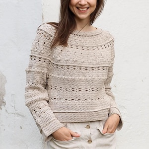 May include: A beige crochet sweater with a round neckline and long sleeves. The sweater features textured patterns and a relaxed fit. The sleeves and body have different stitch patterns, creating visual interest. The garment is made of a soft, neutral-toned yarn.