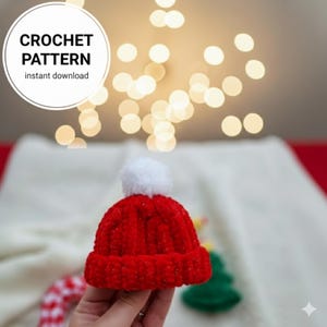 May include: A miniature red crocheted hat with a white pom-pom. The image includes the text "CROCHET PATTERN instant download" in a white circle. The background is softly lit with out-of-focus lights, suggesting a festive theme.