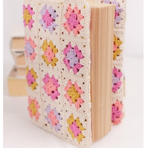 Puede incluir: Un libro envuelto en una cubierta de ganchillo color crema. La cubierta presenta un patrón repetitivo de cuadrados coloridos en rosa, amarillo y lavanda. Las páginas del libro son visibles en el lado derecho.