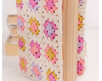 Puede incluir: Un libro envuelto en una cubierta de ganchillo color crema. La cubierta presenta un patrón repetitivo de cuadrados coloridos en rosa, amarillo y lavanda. Las páginas del libro son visibles en el lado derecho.