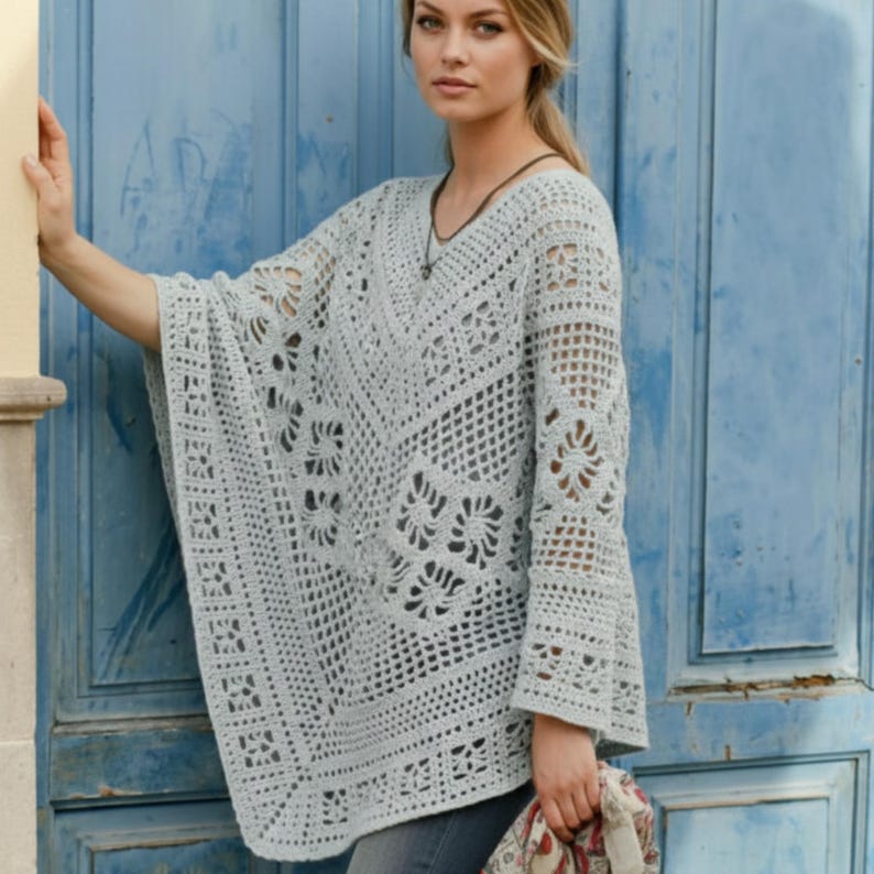 Op de afbeelding: Een lichtgrijze, open geweven gehaakte poncho met een V-hals en klokmouwen. De poncho heeft ingewikkelde bloemen- en geometrische patronen. De vrouw draagt een ketting en houdt een sjaal met patroon vast.