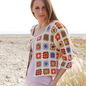 Könnte beinhalten: Cremefarbene gehäkelte Strickjacke mit quadratischem Ausschnitt und Dreiviertelärmeln. Die Strickjacke hat bunte quadratische Motive in Rosa, Orange, Blau und Grün. Das Kleidungsstück ist vorne offen und scheint handgefertigt zu sein.