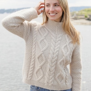 May include: A beige long-sleeve sweater featuring a high mock neck collar, a textured cable knit pattern with vertical braids and diamond shapes on the front, and ribbed cuffs and hem, paired with blue denim jeans.