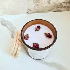 Op de afbeelding: Een geurkaars in een witte en bruine container met een houten deksel. De kaars heeft een witte wasbasis met vier gedroogde rozenknoppen. De container heeft een decoratief etiket met script.