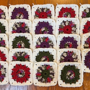 May include: A collection of twenty-four square crochet motifs arranged in a grid. Each square features a central floral pattern in shades of red, purple, and olive green, framed by a white border. The motifs are crafted with yarn.