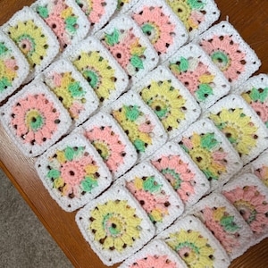 May include: A collection of handmade crochet squares in a variety of pastel colors, including pink, yellow, and green, with a white border. The squares are arranged closely together, showcasing a floral design.