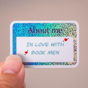 May include: A rectangular sticker with a blue glitter border and the text "About me" in dark blue. The sticker also says "IN LOVE WITH BOOK MEN" with small red heart accents. The sticker is held by a person.