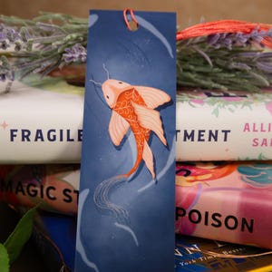 May include: A blue bookmark featuring an illustration of a koi fish in shades of orange and white. The bookmark has a small orange tassel at the top and is placed in front of a stack of books. The words "FRAGILE" and "POISON" are visible on the book spines.